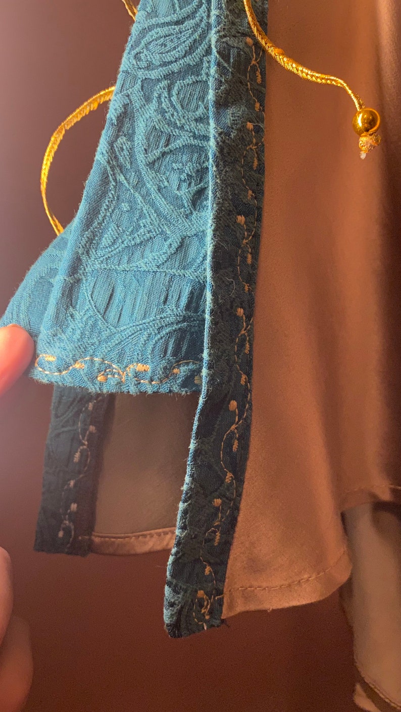 May include: A teal blue fabric with a gold trim and a gold cord with a gold bead at the end. The fabric has a textured pattern.