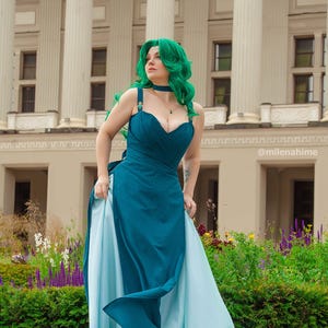 Puede incluir: Un vestido verde azulado y azul claro con escote coraz&oacute;n y falda fluida. La persona de la imagen tiene el pelo verde y una gargantilla a juego. El fondo presenta un edificio con columnas y un jard&iacute;n.
