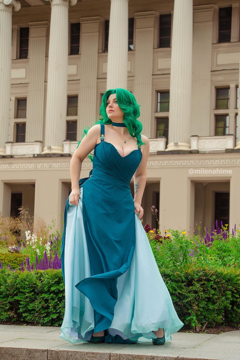 Puede incluir: Un vestido verde azulado y azul claro con escote coraz&oacute;n y falda fluida. La persona de la imagen tiene el pelo verde y una gargantilla a juego. El fondo presenta un edificio con columnas y un jard&iacute;n.