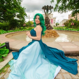 Puede incluir: Un vestido verde azulado y azul claro con una falda fluida. El vestido presenta un cuello halter y lo lleva una persona con cabello verde. El fondo incluye una fuente y &aacute;rboles.