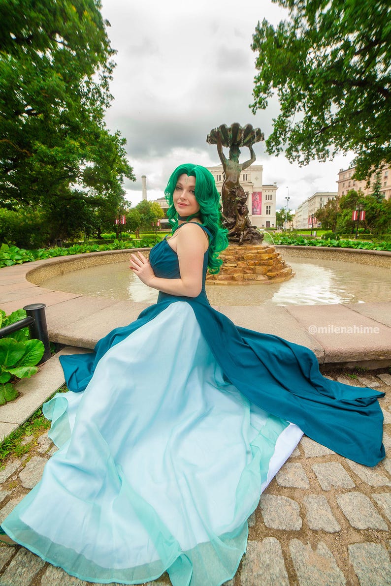 Puede incluir: Un vestido verde azulado y azul claro con una falda fluida. El vestido presenta un cuello halter y lo lleva una persona con cabello verde. El fondo incluye una fuente y &aacute;rboles.