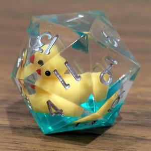 Puede incluir: Un dado transparente de múltiples caras con números plateados, que encierra un patito de goma amarillo. El dado tiene detalles turquesa y está sobre una superficie de madera. Un artículo único para juegos de mesa.