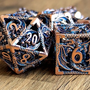 Puede incluir: Un conjunto de dados de seis caras con un diseño de dragón. Los dados están hechos de metal y tienen un acabado de cobre con acentos azules. Los números en los dados son visibles y van del 1 al 6.