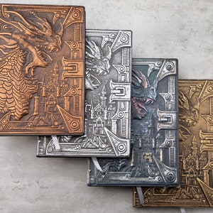 Puede incluir: Cuatro diarios encuadernados en cuero con diseños de dragones y castillos en relieve. Los diarios son en tonos cobre, plata y oro.