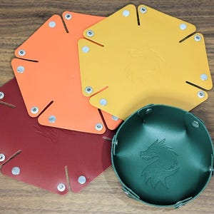 May include: Three flat, hexagonal-shaped trays in orange, yellow, and dark red, alongside one assembled dark green bowl. Each piece features silver metal snaps at the corners and a debossed dragon silhouette logo centered on the surface.