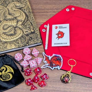 Puede incluir: Un conjunto de accesorios para juegos de mesa, que incluye un diario de dragón dorado en relieve, dados rojos, una bandeja para dados, una bolsa para dados y un llavero. La imagen también incluye un lápiz y una pegatina con el texto "Dice Dragons".