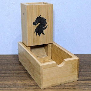 May include: A light brown bamboo dice tower featuring a vertical rectangular shaft with an engraved black dragon silhouette on the front face and an attached open tray at the base for catching dice as they roll through the internal ramp structure.