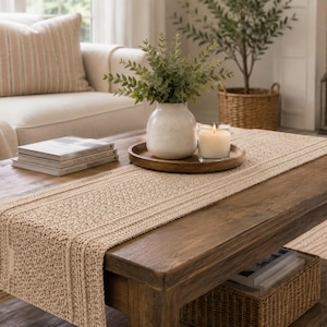 May include: A dark wood coffee table with a woven beige table runner. A white vase with greenery, a wooden tray with a lit candle, and a stack of books are on the table. A beige sofa and a wicker basket are in the background.