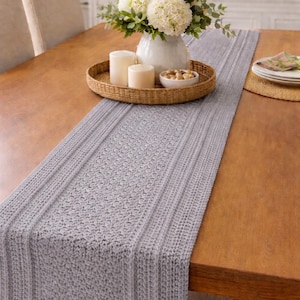 May include: A gray knitted table runner with a textured pattern, placed on a wooden table. A woven tray holds white candles, a white vase with flowers, and a small bowl of nuts. The table is set for a meal.