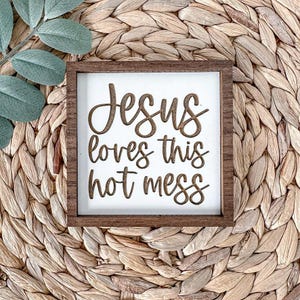 May include: A white wooden sign with brown lettering that reads "Jesus loves this hot mess". The sign is framed in a dark brown wood frame and is sitting on a woven straw mat.