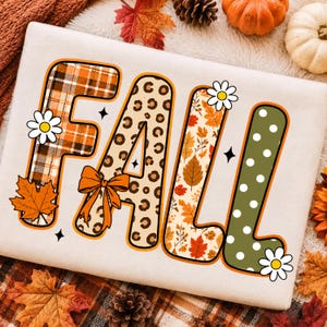 May include: A white sweatshirt featuring the word FALL in large, stylized letters. The F has an orange plaid pattern, the A has a leopard print with an orange bow, the L has an autumn leaf pattern, and the second L is olive green with white polka dots.