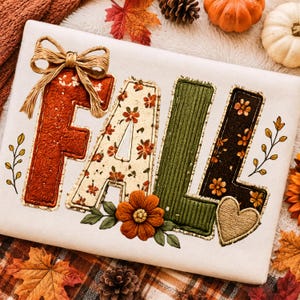 May include: A white sweatshirt featuring the word FALL in large, textured letters. The F is rust-colored with a tan bow, the A has a floral pattern, the L is ribbed green, and the second L is black with floral accents and a small tan heart.