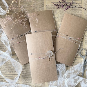 May include: Four brown paper envelopes with white string and small pink and gray embellishments. The envelope in the center has a gold paper clip and a white paper flower.