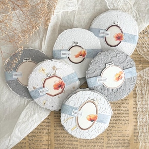 May include: A collection of round, textured coasters in shades of white and gray. Each coaster features a floral design within a decorative frame and a blue ribbon. The coasters have unique patterns and are ideal for home decor.