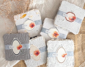 May include: A collection of handmade paper blocks, each wrapped with a light blue ribbon and adorned with a floral sticker inside an embroidery hoop design. The blocks feature embossed patterns and textures in shades of white and gray.