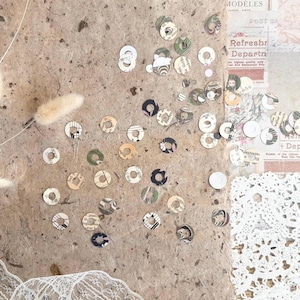 May include: A collection of small, circular paper embellishments with various patterns and colors, including floral, musical notes, and abstract designs. The pieces are scattered on a textured, light brown surface, with hints of lace and vintage ephemera.