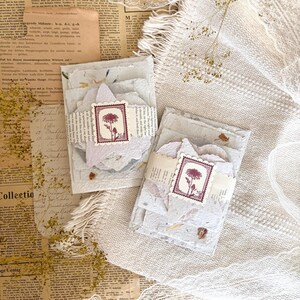 May include: Two stacks of handmade off-white paper sheets with deckled edges, each wrapped in a strip of vintage-style printed text paper. A square stamp featuring a dark red botanical illustration of a flower is centred on each paper band.