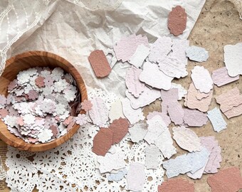 May include: A bowl filled with small, pink and white paper flower confetti. Scattered around the bowl are many small, white, pink, and brown paper tags with scalloped edges.