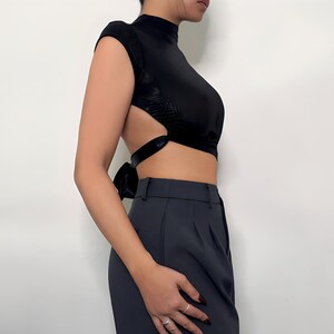 May include: A black mock neck crop top with a tie detail in the back. The top has a cut-out design on the sides. The model is wearing the top with black pants.