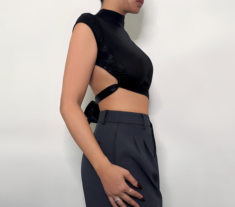 May include: A black mock neck crop top with a tie detail in the back. The top has a cut-out design on the sides. The model is wearing the top with black pants.
