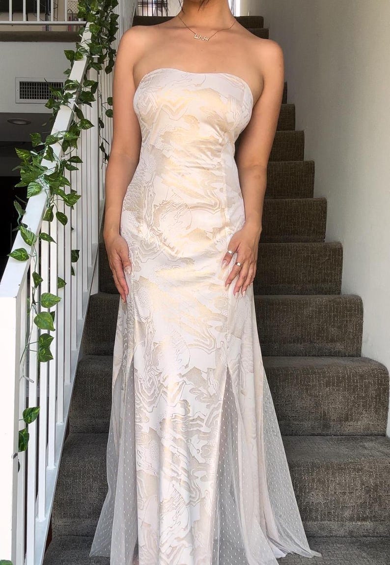 May include: A white strapless gown with a floral pattern and a sheer overlay. The gown has a fitted bodice and a flowing skirt.