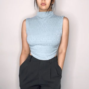 May include: A light blue sleeveless top with a mock neck and a textured pattern. The top is fitted and cropped, and the fabric has a slight sheen.