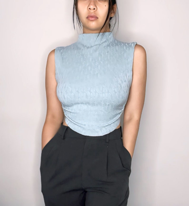 May include: A light blue sleeveless top with a mock neck and a textured pattern. The top is fitted and cropped, and the fabric has a slight sheen.
