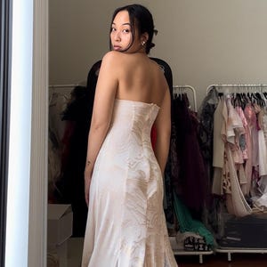 May include: A woman wearing a white strapless gown with a floral pattern and a sheer overlay with white polka dots. The gown has a fitted bodice and a flowing skirt.