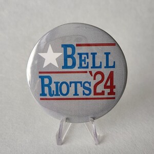 May include: A silver button with a red, white, and blue design. The button says "BELL RIOTS'24" with a white star above the word "BELL".