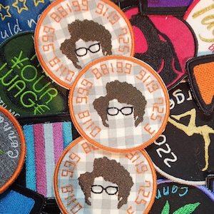 Puede incluir: Tres parches redondos bordados con una imagen de dibujos animados de una persona con el pelo rizado y gafas. Los parches son blancos con un patrón de cuadros blanco y negro y un borde naranja. El texto alrededor de la imagen es "0118 999 88199 9119 725 3".