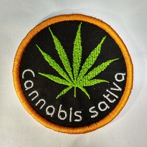 May include: A round, orange-bordered patch with a black background and a green marijuana leaf in the center. The text "Cannabis sativa" is written in white around the leaf.