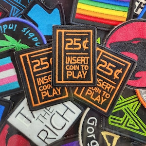 Könnte beinhalten: Eine Sammlung bunter gestickter Patches mit verschiedenen Designs, darunter eine Regenbogenflagge, ein Friedenszeichen, ein Marihuanablatt und ein Retro-Arcade-Spiel-Einsatz mit dem Text "25¢ INSERT COIN TO PLAY".