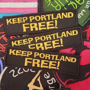 May include: Close-up of several embroidered patches. Three black patches with the text "KEEP PORTLAND FREE!" in yellow are visible. Other patches with various designs and text are in the background, including a transgender pride flag.