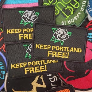 May include: Black rectangular patches with the text "KEEP PORTLAND FREE!" in yellow, accompanied by a white rose with green leaves. Other patches with various designs and text are visible in the background.
