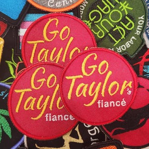 Puede incluir: Una colección de parches bordados. Tres parches circulares rojos con el texto "Go Taylor" en escritura amarilla son el punto focal. La palabra "fiancé" está debajo del nombre. Se ven otros parches con varios diseños y texto.