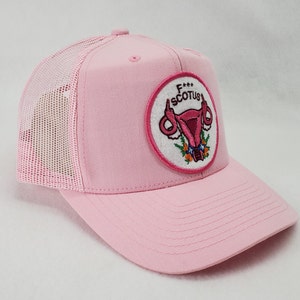 May include: Pink trucker hat with a white patch featuring a pink uterus with a floral design and the text "F*** SCOTUS".