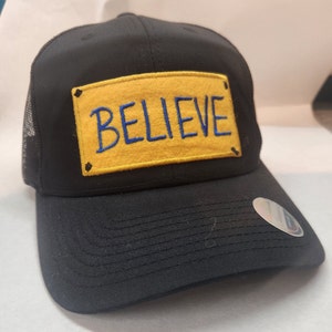 May include: A black trucker hat with a yellow patch that says "BELIEVE" in blue embroidery.
