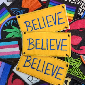 Può includere: Una collezione di toppe ricamate con vari disegni. Tre toppe rettangolari gialle con la parola "BELIEVE" in blu sono in evidenza. Altre toppe presentano strisce arcobaleno, testo e disegni grafici in vari colori.