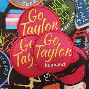 Puede incluir: Una colección de parches bordados con varios diseños. Varios parches circulares rojos presentan el texto "Go Taylor's husband" en escritura amarilla. Otros parches incluyen texto y gráficos, como una hoja de cannabis y un arcoíris.