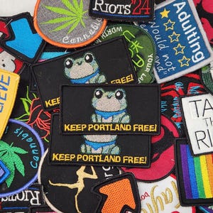 May include: A collection of embroidered patches with various designs and text. Notable patches include a frog with "Keep Portland Free!", a cannabis leaf, "Tax the Rich", and "Believe". The patches are in various colors and shapes.