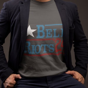 May include: A gray t-shirt with a blue and red graphic that says "BELIEVE" and "RIOTS 24" with a white star.