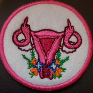 May include: Pink embroidered patch of a uterus with two middle fingers raised, surrounded by green leaves and orange and blue flowers.