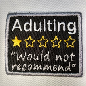 Op de afbeelding: Geborduurde patch met het woord "Adulting" in wit, boven vijf gele sterren. Onder de sterren is de zin "Would not recommend" in wit geborduurd. De patch heeft een zwarte achtergrond met een grijze rand.