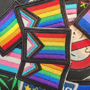 May include: A close-up of several embroidered patches featuring various pride flags, including the rainbow flag, the transgender flag, and the bisexual flag. The patches are all sewn onto a dark fabric background.