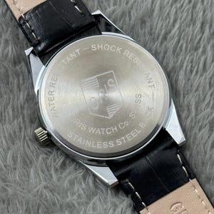 K&ouml;nnte beinhalten: Eine silberfarbene Uhr mit schwarzem Lederarmband. Auf der R&uuml;ckseite der Uhr steht "WATER RESISTANT" und "SHOCK RESISTANT". Die Uhr zeigt "ORIS WATCH CO. SWISS STAINLESS STEEL BACK". Das Armband ist aus schwarzem Leder.
