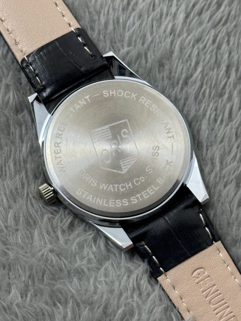 K&ouml;nnte beinhalten: Eine silberfarbene Uhr mit schwarzem Lederarmband. Auf der R&uuml;ckseite der Uhr steht "WATER RESISTANT" und "SHOCK RESISTANT". Die Uhr zeigt "ORIS WATCH CO. SWISS STAINLESS STEEL BACK". Das Armband ist aus schwarzem Leder.