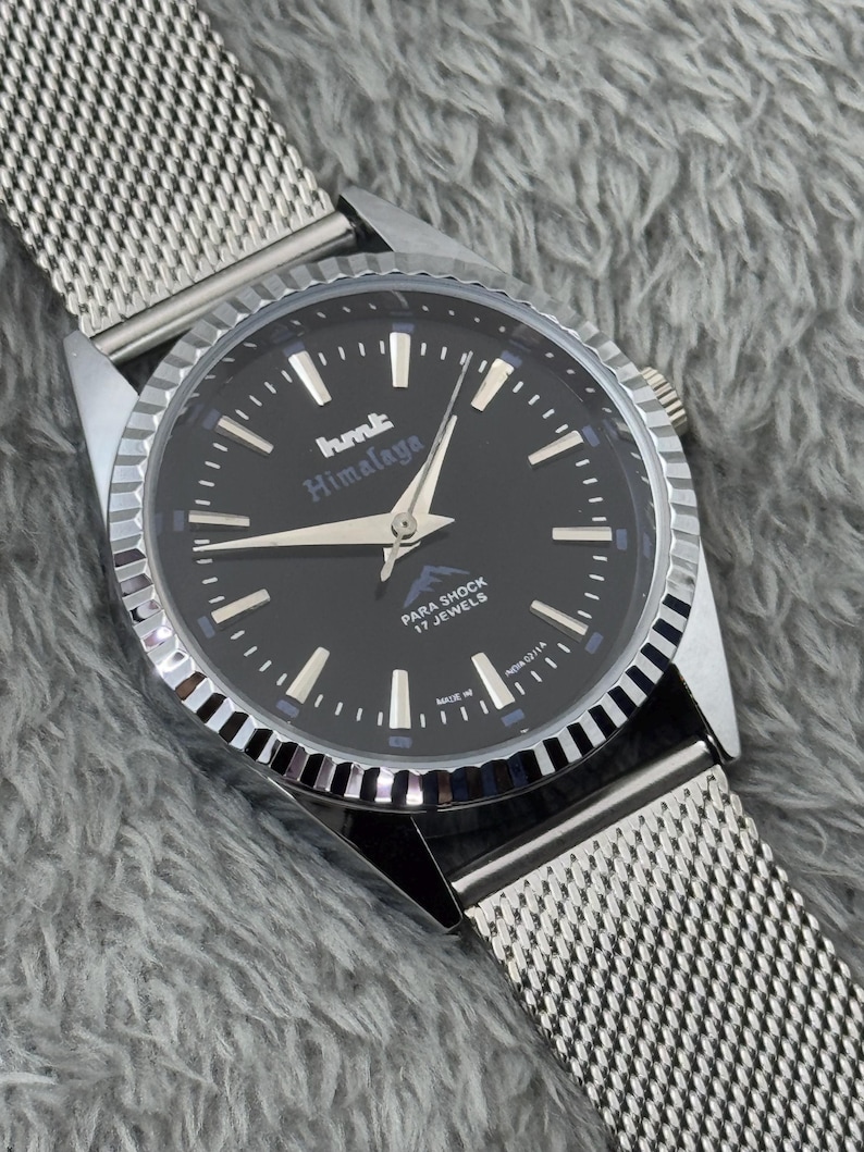 Puede incluir: Un reloj de pulsera plateado con esfera negra y correa de malla. La esfera del reloj presenta marcadores de hora y agujas blancas, y el texto "hmt Himalaya". El reloj tambi&eacute;n tiene el texto "PARA SHOCK 17 JEWELS".