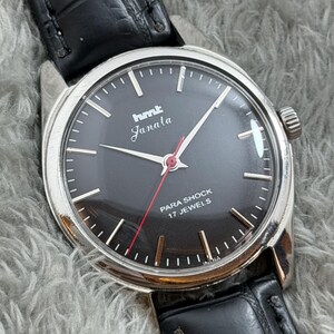 Op de afbeelding: Een vintage HMT Janala polshorloge met een zwarte wijzerplaat, zilveren kast en zwarte leren band. Het horloge heeft witte uurmarkeringen, zilveren wijzers en een rode secondewijzer. De wijzerplaat leest "PARA SHOCK 17 JEWELS" en "INDIA".