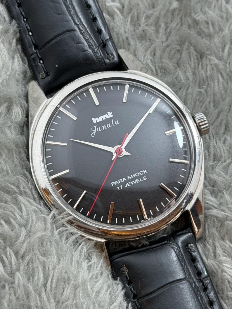 Op de afbeelding: Een vintage HMT Janala polshorloge met een zwarte wijzerplaat, zilveren kast en zwarte leren band. Het horloge heeft witte uurmarkeringen, zilveren wijzers en een rode secondewijzer. De wijzerplaat leest "PARA SHOCK 17 JEWELS" en "INDIA".