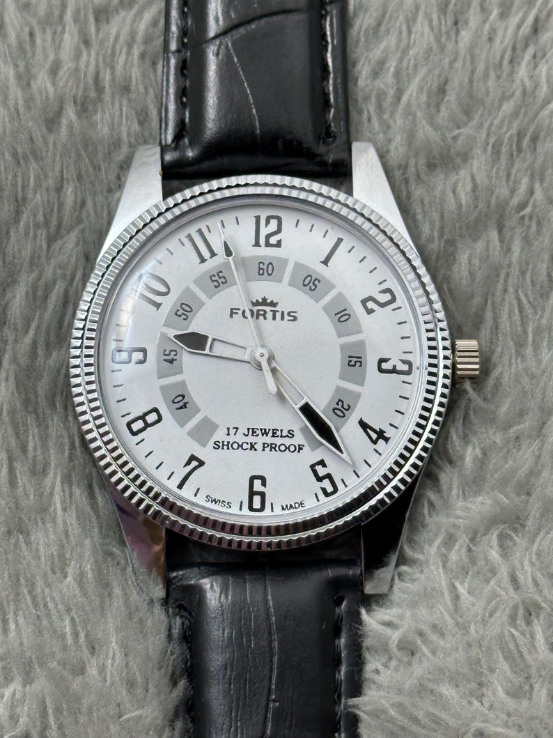 K&ouml;nnte beinhalten: Silberfarbene Fortis-Uhr mit wei&szlig;em Zifferblatt, schwarzen Zeigern und schwarzem Lederarmband. Das Zifferblatt zeigt das Fortis-Logo, die Aufschrift "17 Jewels Shock Proof" und die Zahlen 1 bis 12. Die Uhr hat eine strukturierte L&uuml;nette.